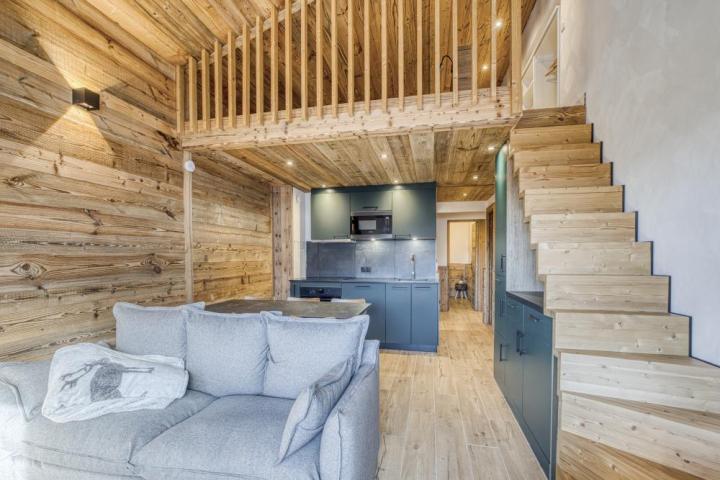 Rénovation intérieure d'un chalet en vieux bois à l'atmosphère authentique et chaleureuse. Studio salon / cuisine avec un canapé gris et des façades de mobiliers bleu-vert. Habillage mural, escalier, mezzanine en vieux bois.