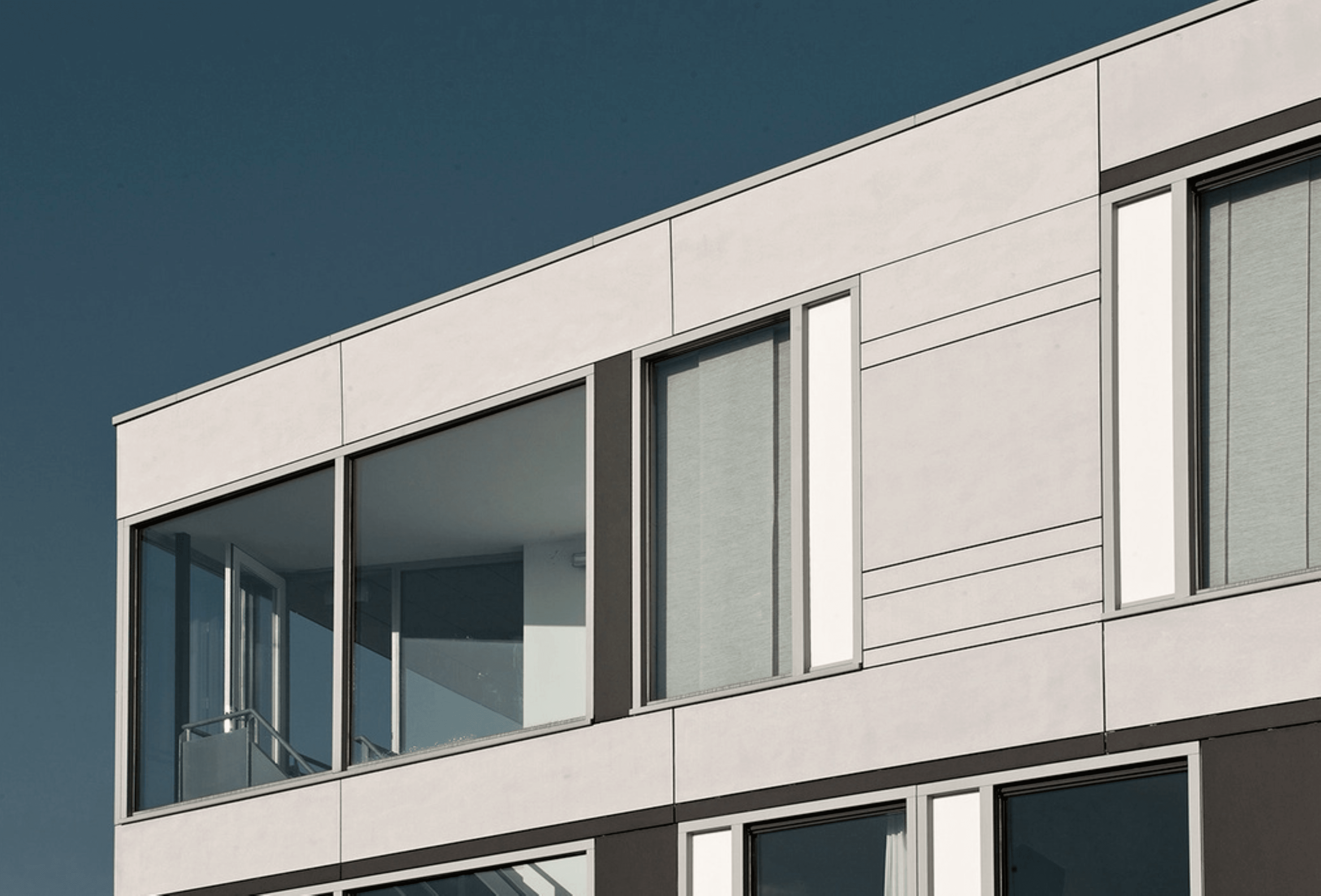 Photo de réalisation. Haut d'un bâtiment avec des panneaux de façade gris clairs. Grandes fenêtres.