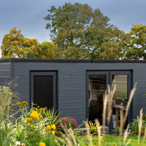 Petite habitation en bardage composite de couleur gris anthracite, avec trois portes vitrées. Elles est entourée d'un jardin en fleurs. 