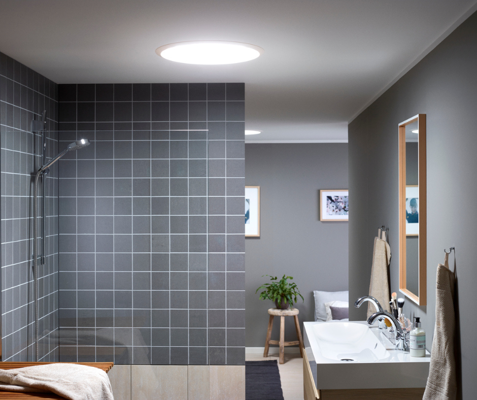 Réalisation d'un conduit de lumière sun tunnel velux au plafond d'une salle de bain. Le puit de lumière illumine la pièce. Le mur de la douche est en carrelage gris foncé. Sur la droite il y a un lavabot avec un miroir. 