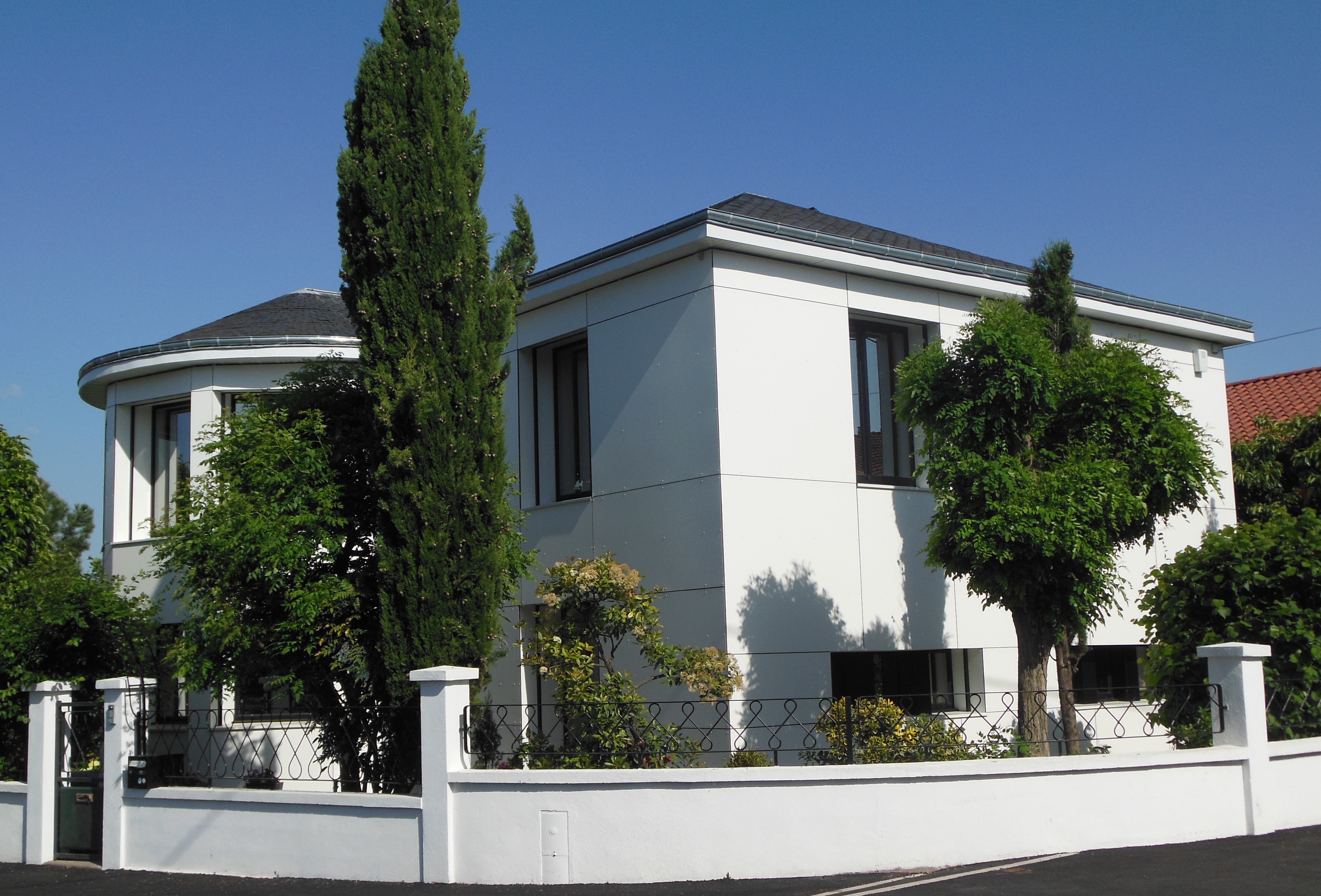 Photo de réalisation. Maison avec des panneaux de façade blancs. Hauts sapins verts autour de la maison. Muret blanc encerclant la maison. 