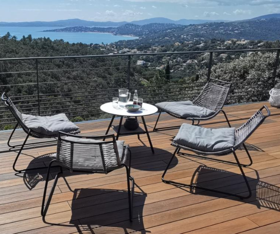 Prise de vue dehors donnant sur un lac et les arbres. Sur la terrasse atmosphère nuances IPE il y a 4 chaises avec un coussin gris sur chacune et une petite table ronde blanch au milieu. 