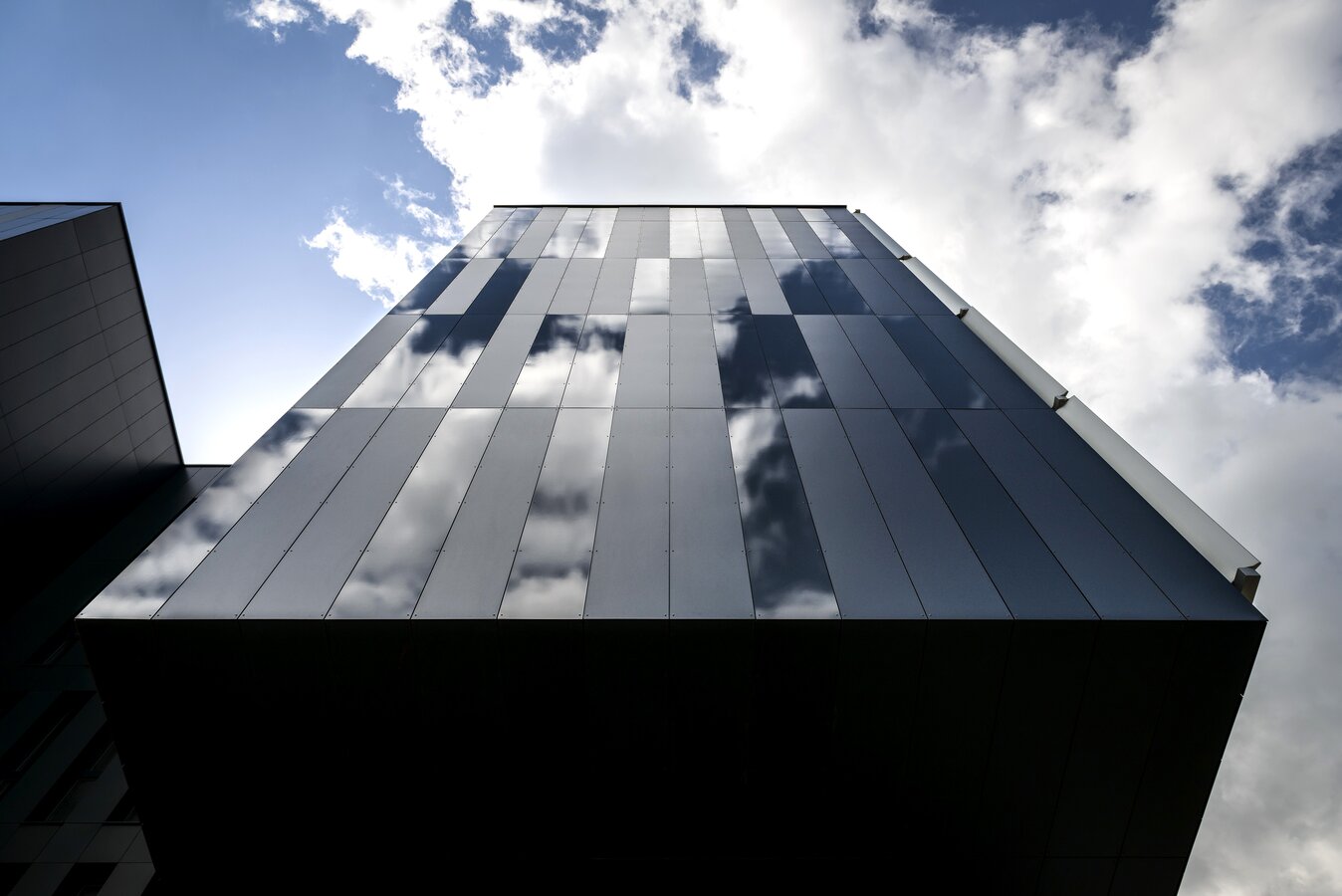 Prise de vue d'un bâtiment en contre-plongée. Panneaux de façades bleus marines et quelques-uns sont en verres. Nuages au-dessus du bâtiment.