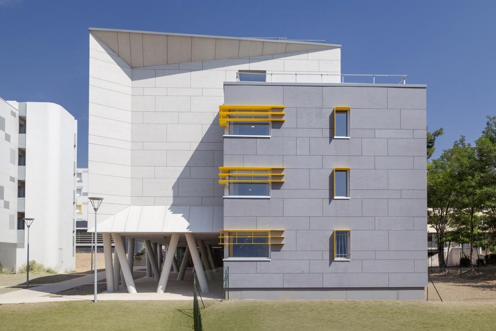 Extérieur bâtiment. Panneaux de façade gris avec des contours de fenêtre jaune. En arrière plan, un bâtiment surélevé, à l'architecture moderne et des panneaux de façade blanc. Au sol pelouse verte. 