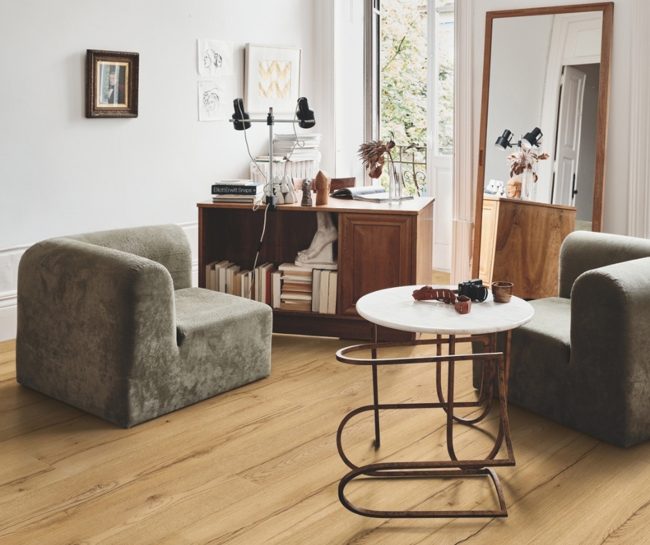 Mise en avant dans un salon du sol stratifié PERGO de couleur marron clair. Dans la pièce il y a une table basse blanche avec les pieds marrons, deux coins de fauteuils gris, un grand miroir et un bureau dans le fond. 