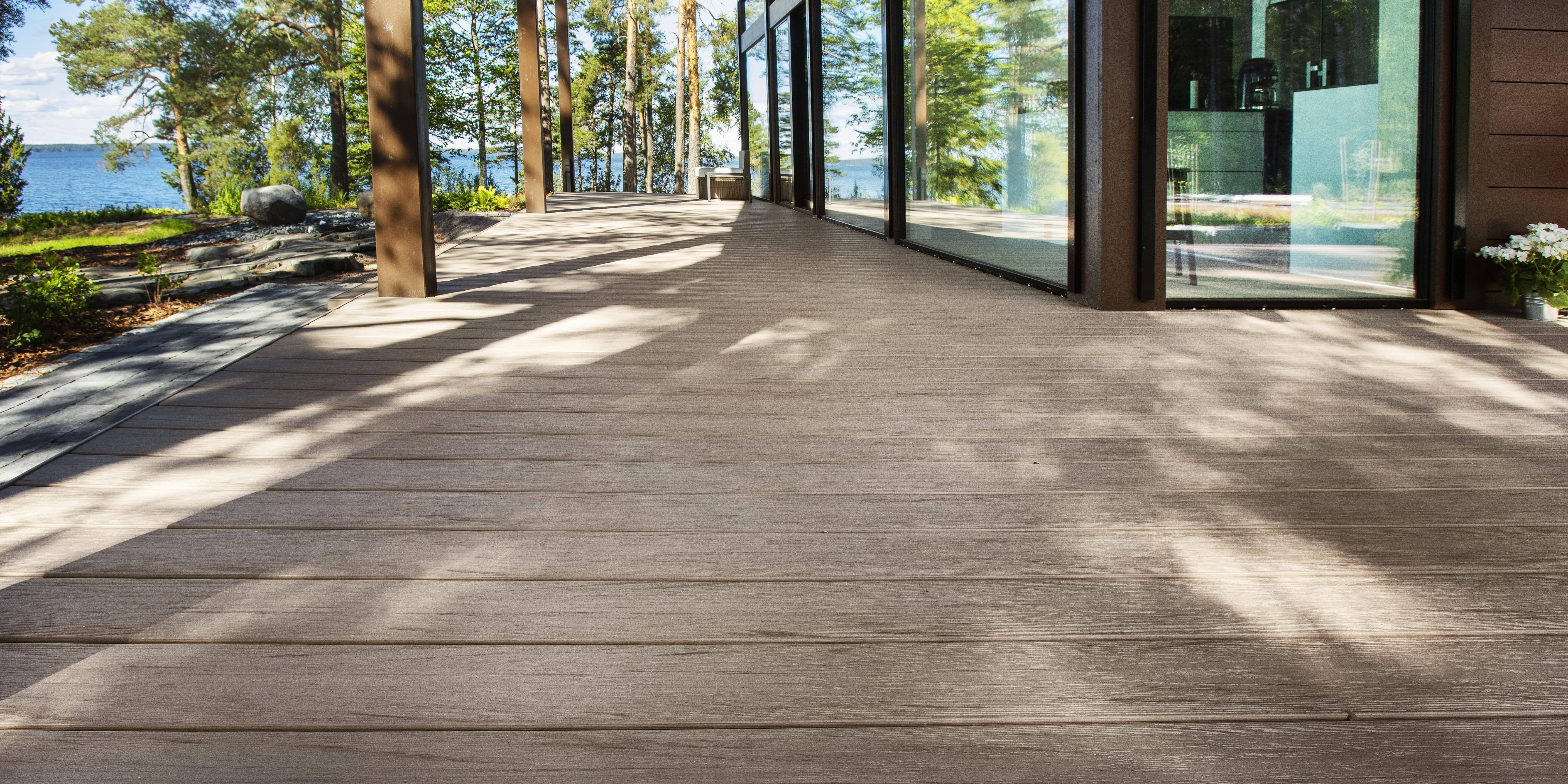 Terrasse extérieure en bois composite marron clair. Baies vitrées de la maison sur le côté droit et arbres sur le côté gauche. Mer bleue en arrière-plan. 