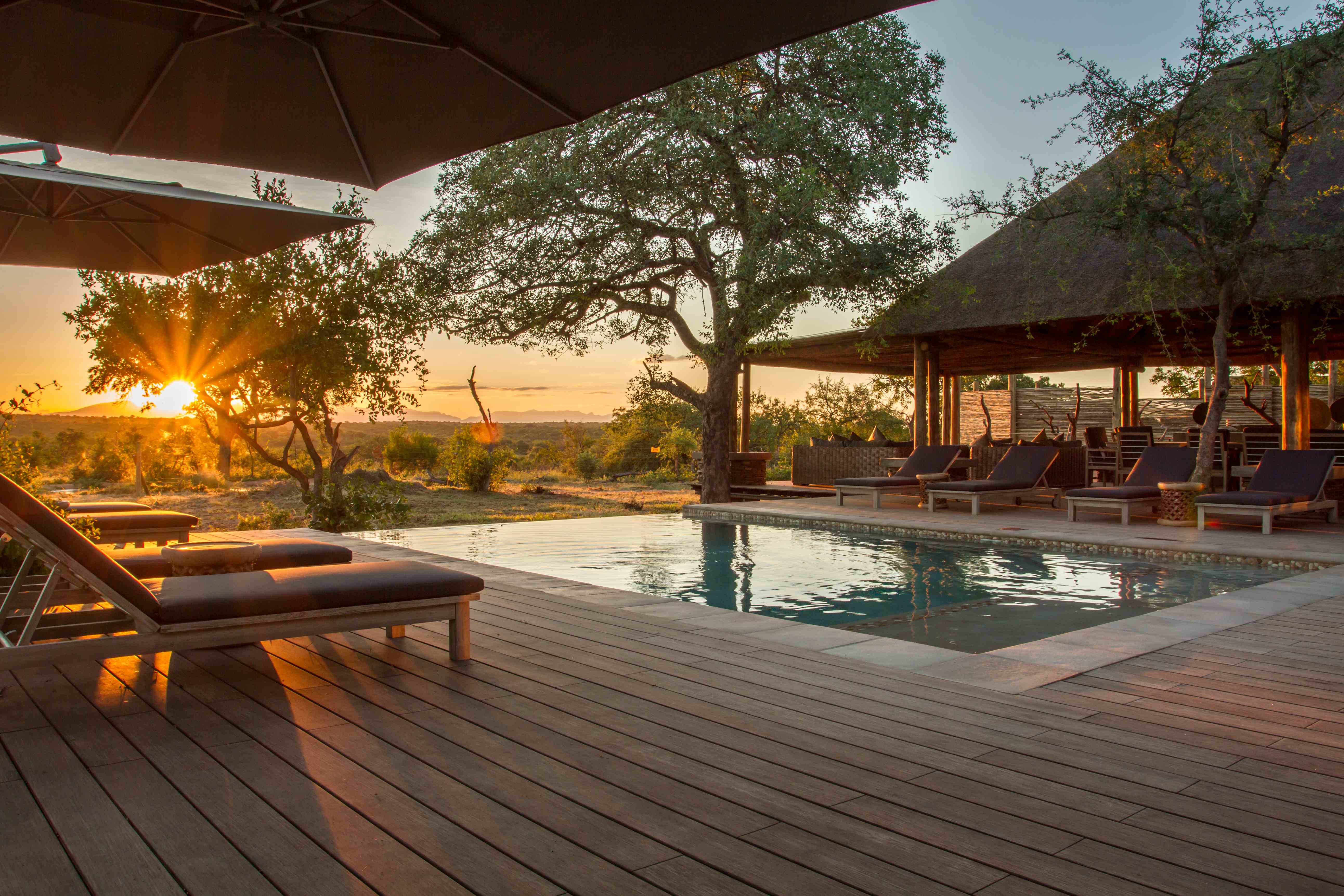 Terrasse en bambou, de couleur brun. Piscine au milieu de la terrasse. Transats, parasols et arbres autour. Le tout est accompagné d'un magnifique couché de soleil.