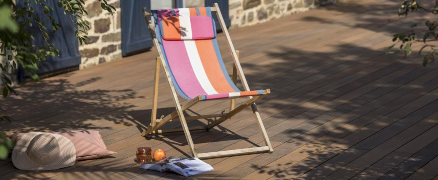 Réalisation de la terrasse composite Atmosphère Nuances IPE de Silvadec. Une chaise de vacance en tissu est présente, de couleur rose, orange blanche et bleu. Sur la gauche au sol il y a un chapeau beige et un coussin. 