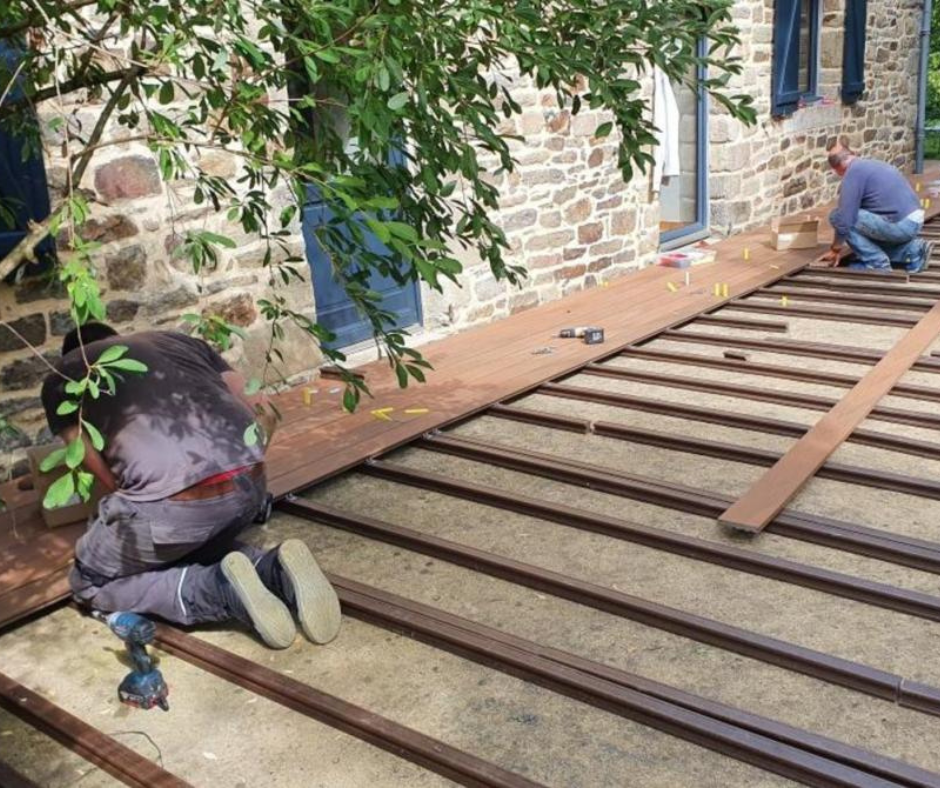 Deux messieurs posent les lames de terrasse. La structure est posée et deux rangées de lames de terrasse sont déjà installées. Elle est réalisée au bord d'une maison en pierre avec des menuiseries bleues. 