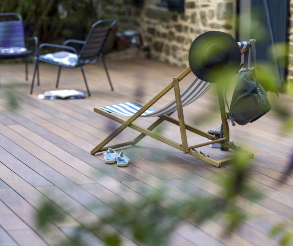 Photo de réalisation d'une terrasse Atmosphère Nuances IPE. Un chaise longue rayée bleu et blanc est présente. Au premier plan des plantes vertes floutées sont là.