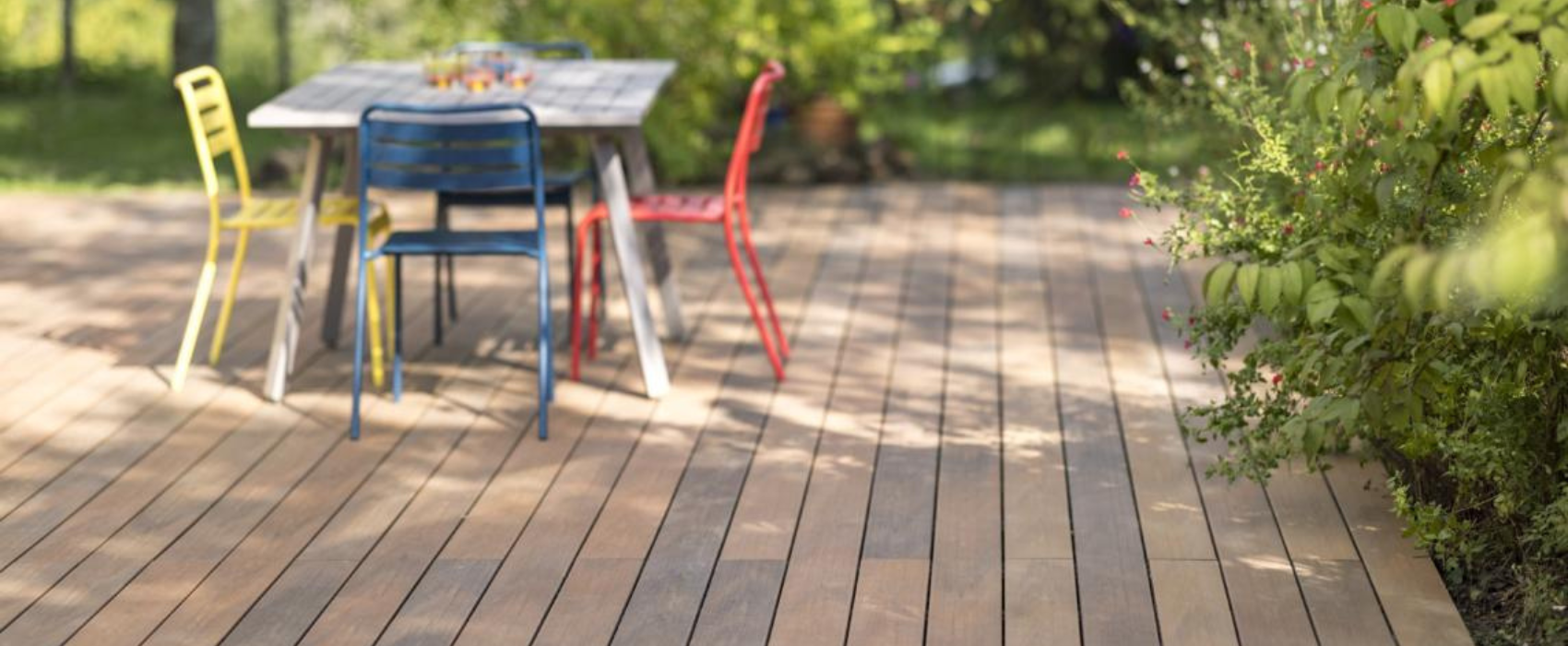 Réalisation d'une terrasse composite Atmosphère Nuances IPE de Silvadec. Des buissons entours la terrasse composite. Au milieu, une table avec trois chaises (jaune, bleu, rouge).