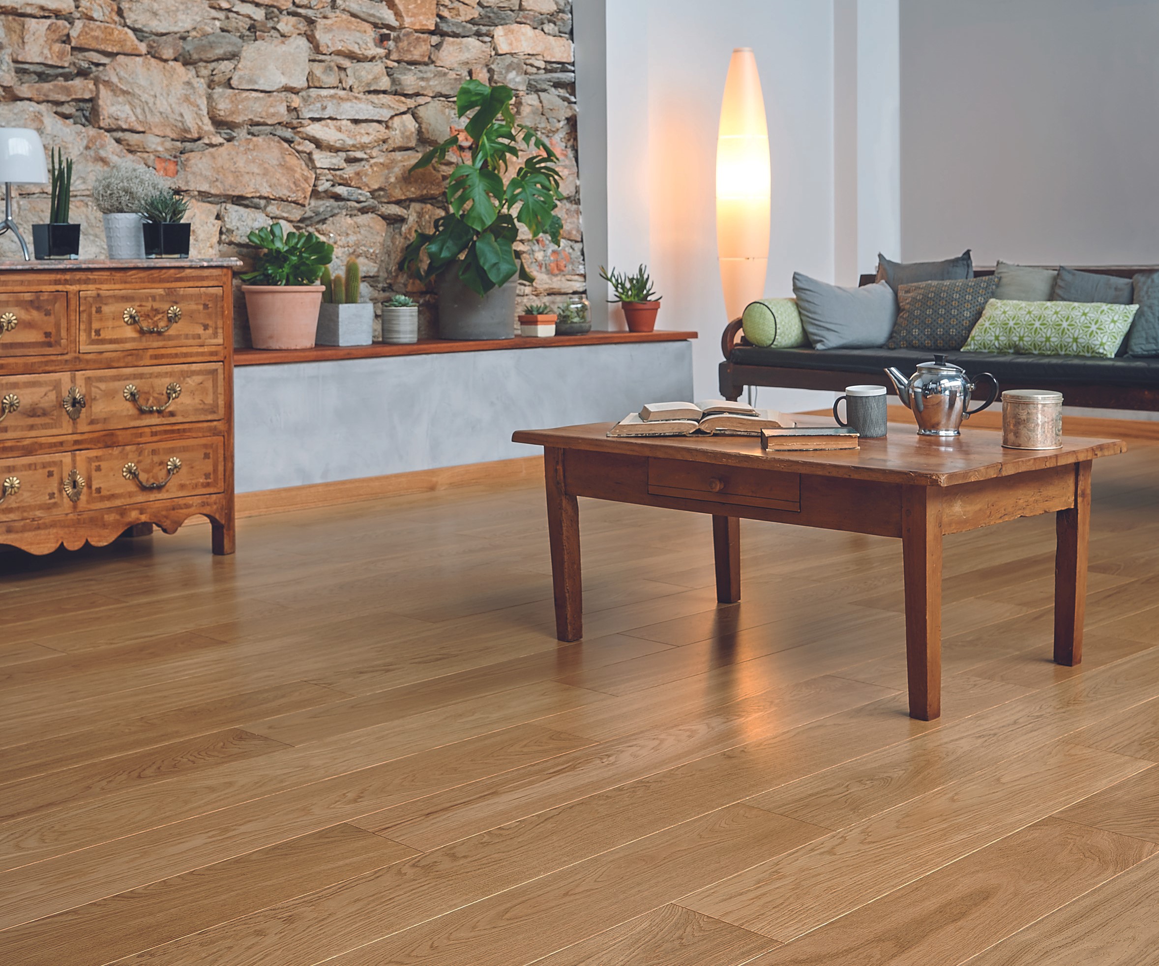 Parquet chêne massif de couleur miel installé dans le salon. La table basse et la commode sont en bois. En arrière-plan il y a un canapé avec des cousins verts dessus, ainsi que des plantes et une lumière à même le sol en guise de décoration.