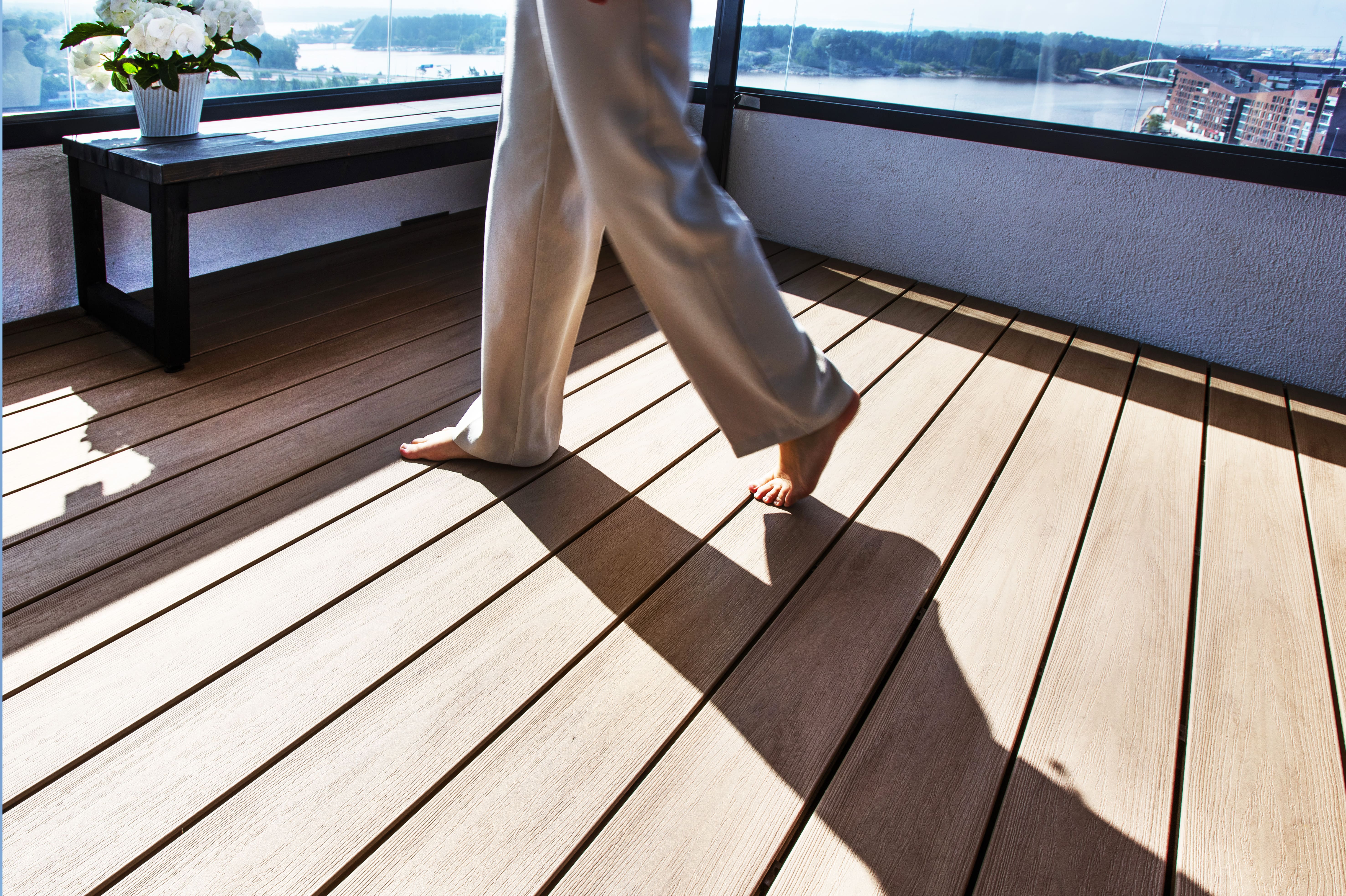 Terrasse de balcon de couleur claire. On voit les jambes d'une personne qui marche pieds nues avec un pantalon blanc. Table sur la terrasse et mer bleue en arrière-plan.